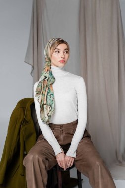 Başörtüsü ve beyaz balıkçı yakalı kadın gri arka planda perdenin yanında oturuyor.