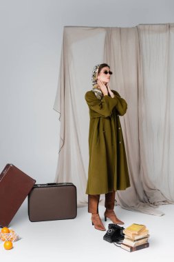 Güneş gözlüklü zarif bir kadın bavulların yanında duruyor ve gri örtülü arka plan kitaplarının yanında klasik bir telefon var.