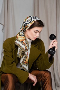 Gri kumaşlı, bulanık arka planda, antika telefon ahizesiyle oturan, desenli mendilli bir kadın.