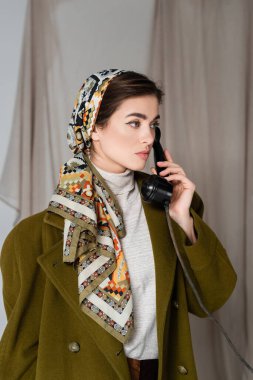 Şablonlu başörtüsü takmış şık bir kadın gri arka plandaki eski moda telefondan arıyor.