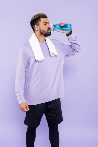 bearded african american man in sweater standing with white towel and drinking water isolated on purple