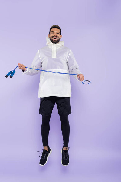 full length of happy african american man in sportswear levitating and holding jumping rope on purple