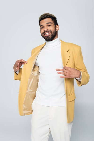 cheerful african american man adjusting yellow blazer isolated on grey 