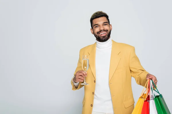 黄色のブレザーとポロネックの幸せなアフリカ系アメリカ人男性グレーで隔離されたシャンパンのショッピングバッグとガラスを保持 — ストック写真