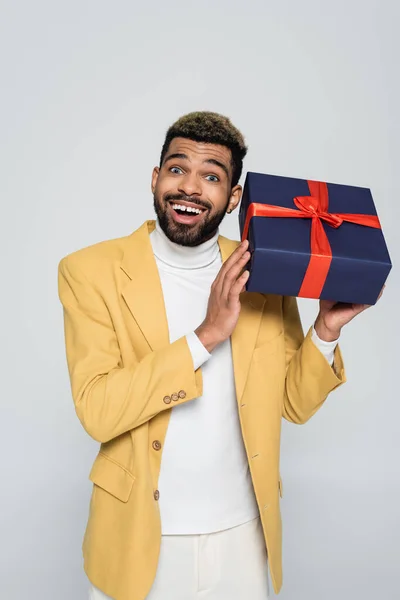 興奮アフリカ系アメリカ人男性で黄色スタイリッシュなブレザー保持ラッププレゼント絶縁上グレー — ストック写真
