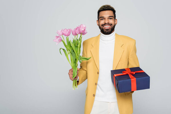 happy african american man in yellow stylish blazer holding wrapped present and tulips isolated on grey 