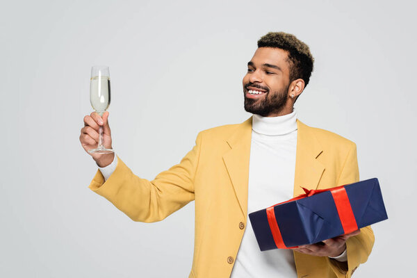 bearded african american man in yellow stylish blazer holding wrapped present and glass of champagne isolated on grey 