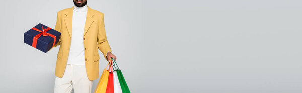 cropped view of bearded african american man in yellow blazer holding present and shopping bags isolated on grey, banner 