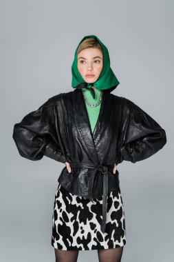 Deri ceketli ve yeşil mendilli, elleri belinde gri renkli poz veren moda bir kadın.