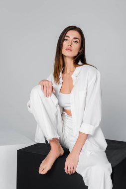 Esmer, çıplak ayaklı, siyah küpün üzerinde oturan ve gri renkli kameraya bakan kadın.