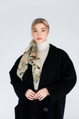 Siyah ceketli güzel bir kadın ve şık bir başörtüsü var. Gri renkli kameraya bakıyor.