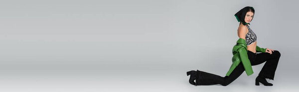 full length of woman in kerchief and black jeans posing on one knee on grey background, banner