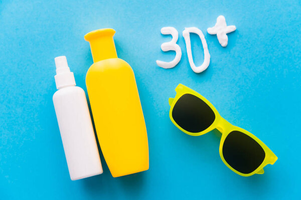 Top view of sunscreens near 30+ signs and sunglasses on blue background 