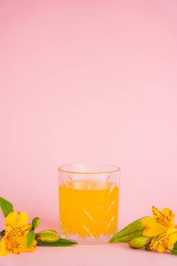 Bir bardak taze narenciye suyu ve pembe arka planda sarı tropik çiçekler.