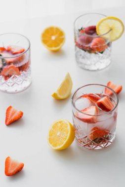 Meyveli tonik bardaklar, doğranmış çilek ve limonların yanında beyaz masanın üstünde.