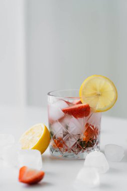 Beyaz yüzeyde kesilmiş limon ve gri arka planda doğranmış çilekli buzlu tonik.
