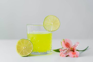 Pembe alstroemeria çiçeği ve dilimlenmiş limonata, gri yüzeyde izole edilmiş beyaz limonata camının yanında.