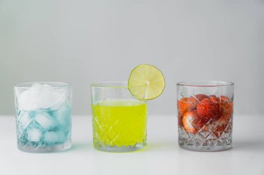 lemonade and strawberry tonic near glass with ice cubes on white surface isolated on grey clipart