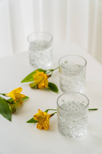 crystal glasses with water near yellow alstroemeria flowers on white tabletop and blurred background 