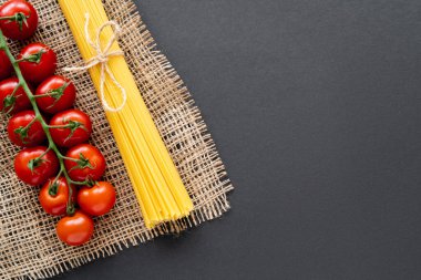 Taze vişneli domates ve siyah arka plandaki çuval bezi üzerinde pişmemiş makarna manzarası. 