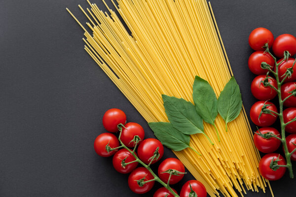Top view of raw spaghetti near cheery tomatoes and basil on black background 