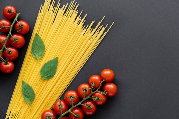 Top view of raw spaghetti near fresh cherry tomatoes and basil on black background 