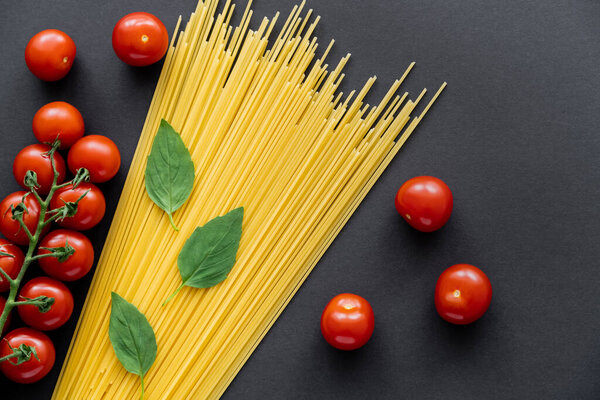 Top view of whole cherry tomatoes near fresh basil and raw spaghetti on black background 