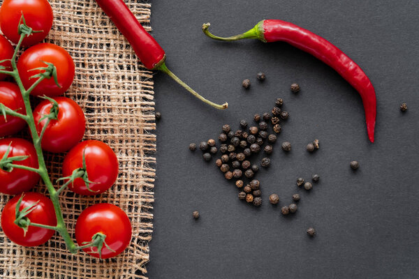 Top view of cherry tomatoes and chili peppers near peppercorns on sackcloth on black background 