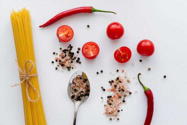 Top view of raw pasta near spices with cherry tomatoes and chili pepper on white background 