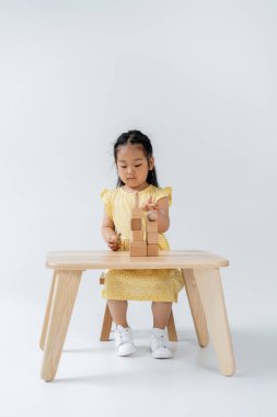 Sarı elbiseli Asyalı anaokulu öğrencisi kız gri üzerinde tahta şekillerle oynuyor.
