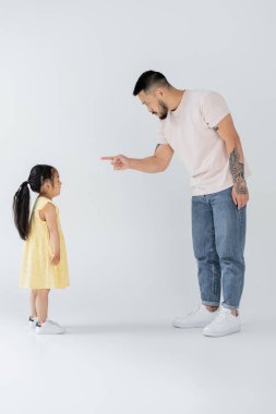 full length of asian father pointing at preschooler daughter while punishing her on grey clipart