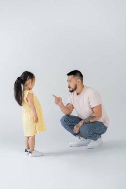 Tam boy Asyalı dövmeli baba, anaokulu kızına işaret ederken onu gri ile cezalandırıyor.