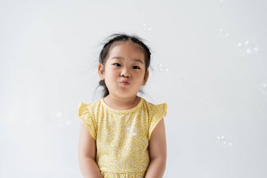 portrait of asian preschooler girl in yellow dress pouting lips near near soap bubbles isolated on grey  clipart