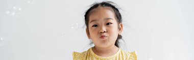 portrait of asian preschooler girl pouting lips near near soap bubbles isolated on grey, banner clipart