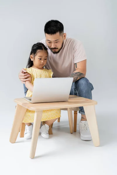 tattooed father and asian daughter looking at laptop on grey