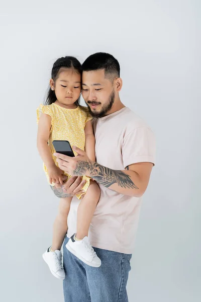 Asiatischer Tätowierter Mann Benutzt Smartphone Und Hält Tochter Arm Isoliert — Stockfoto