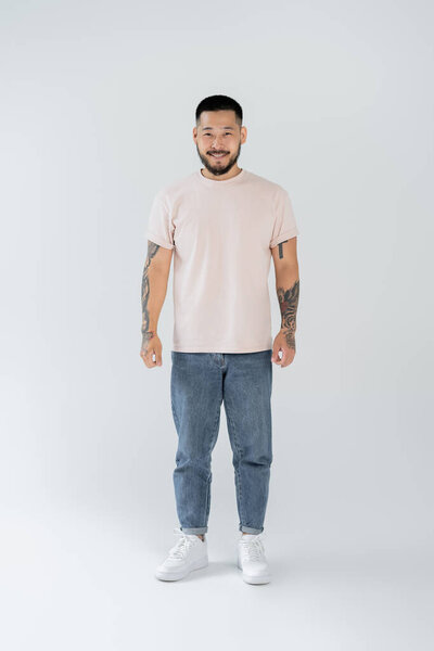 full length of happy asian man with beard standing in casual clothing on grey