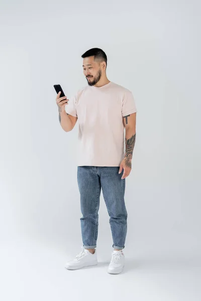 Full Length Smiling Asian Man Tattoos Using Smartphone Grey — Stock Photo, Image