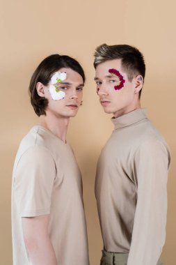 Young homosexual couple with petals on faces looking at camera isolated on beige  clipart