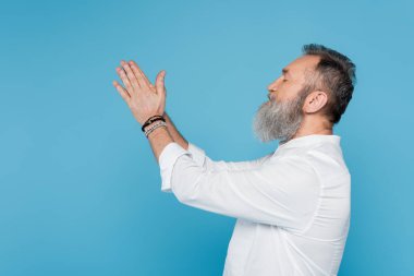 Beyaz gömlekli guru akıl hocasının yan görünüşü, dua eden ellerle meditasyon yaparken mavi üzerine izole edilmiş.