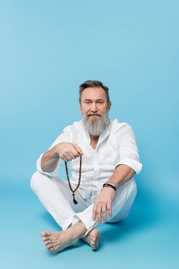 Bacak bacak üstüne atmış sakallı, yalınayak bir adam mavi arka planda mala boncukları tutuyor.