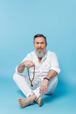 bearded barefoot man sitting with crossed legs and holding mala beads on blue background clipart
