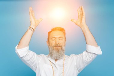 master guru meditating with closed eyes and raised hands near shining aura isolated on blue clipart