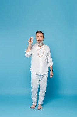 Mavi arka planda selenit kristali tutan çıplak ayaklı yoga öğretmeni.