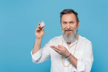 Sakallı usta guru selenit kristali gösteriyor ve mavi üzerine izole edilmiş kameraya bakıyor.