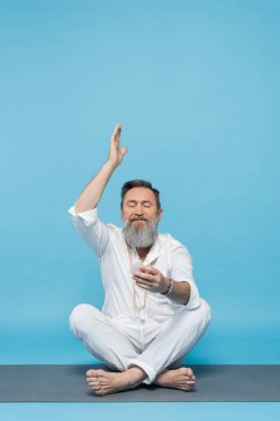 senior man in white clothes meditating with selenite crystal in easy pose with raised hand on blue clipart