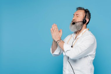 Kulaklık takmış usta guru, gözleri kapalı meditasyon yapıyor ve dua eden eller mavi üzerine izole edilmiş.