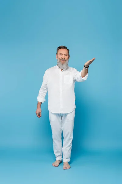 full length of bearded barefoot man in white clothes pointing with hand on blue