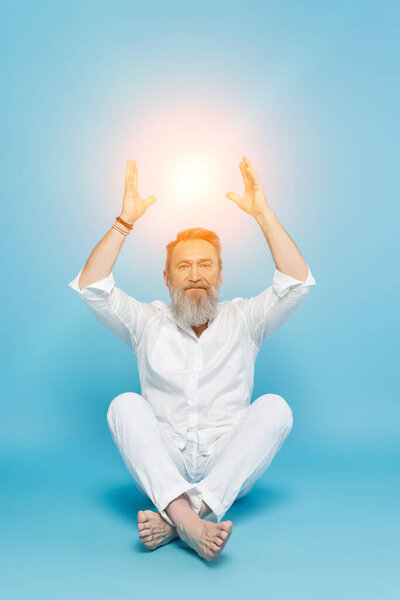 bearded man in white clothes meditating near shining aura on blue background