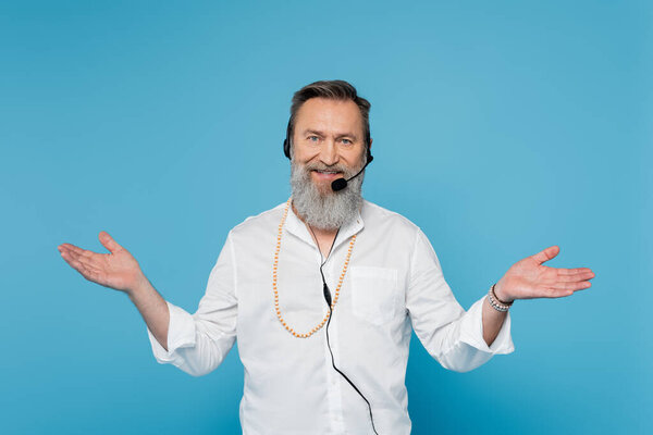 улыбающийся мотивационный тренер в наушниках, стоящий с распростертыми объятиями, изолированный на голубом